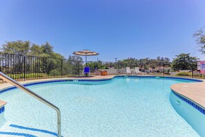 A clear swimming pool with a blue tiled edge, surrounded by a metal fence, and a shaded area with a table and chairs.