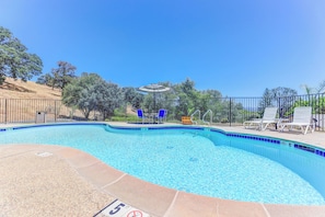 A clear swimming pool with a blue tiled edge, surrounded by a fence, and a covered area with chairs and a table.