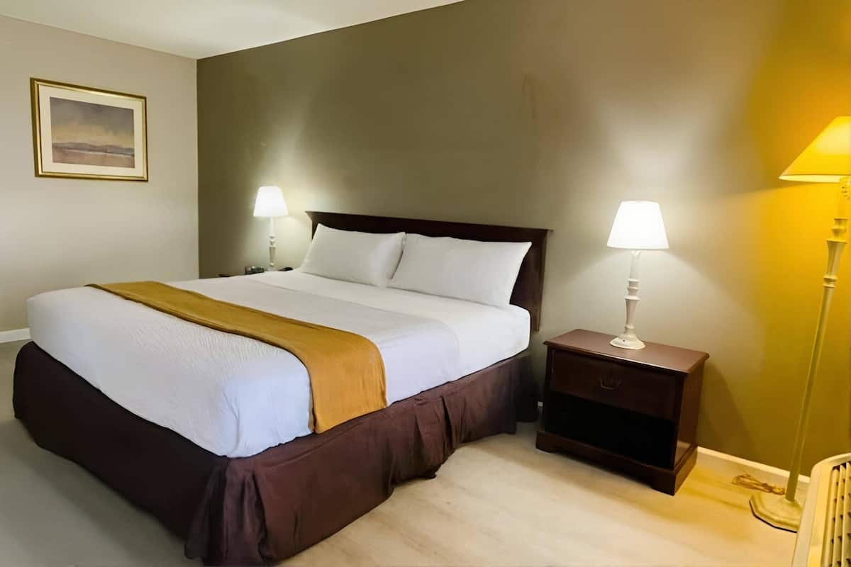 A neatly made bed with white linens and a brown accent, flanked by two bedside tables with lamps, in a room with a neutral color scheme and a framed picture on the wall.
