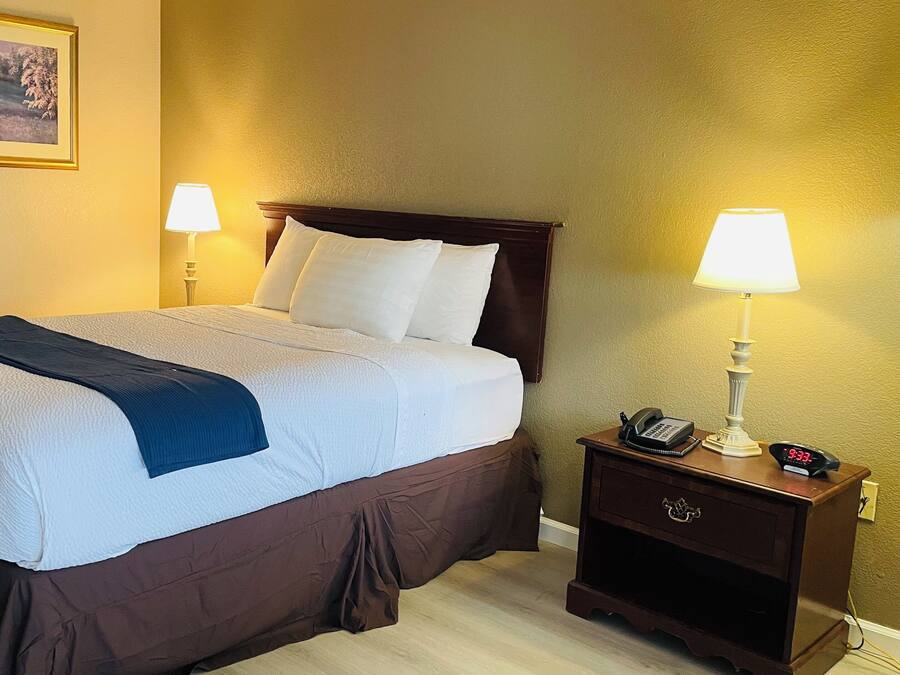 A hotel room with a large bed, two bedside lamps, a nightstand with a phone and a clock, and a framed picture on the wall.