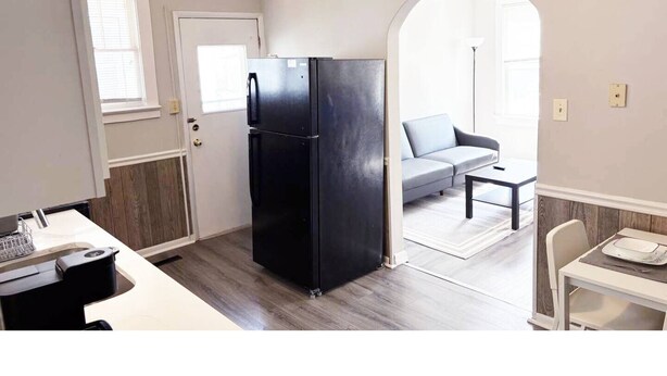 A compact kitchen with a black refrigerator, a dining area with a white table and chairs, and a gray sofa in the adjacent living room.