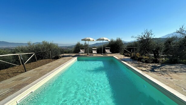 A rectangular swimming pool with clear water, surrounded by a paved deck and a fence. There are two umbrellas and chairs in the background. - Self-contained apartment with independent access in Farmhouse. (Toscana)
