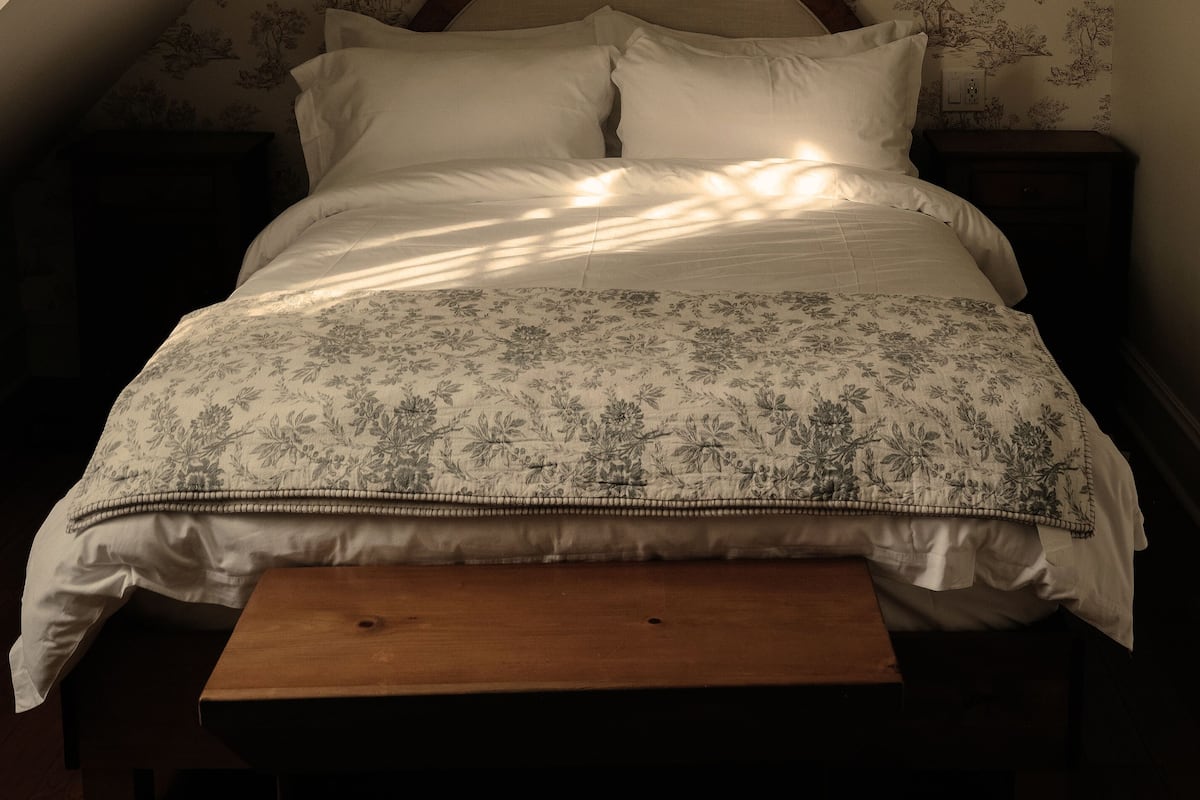 Une chambre avec un plafond en pente, une tête de lit en bois, un lit avec une literie à motifs floraux et un luminaire suspendu.