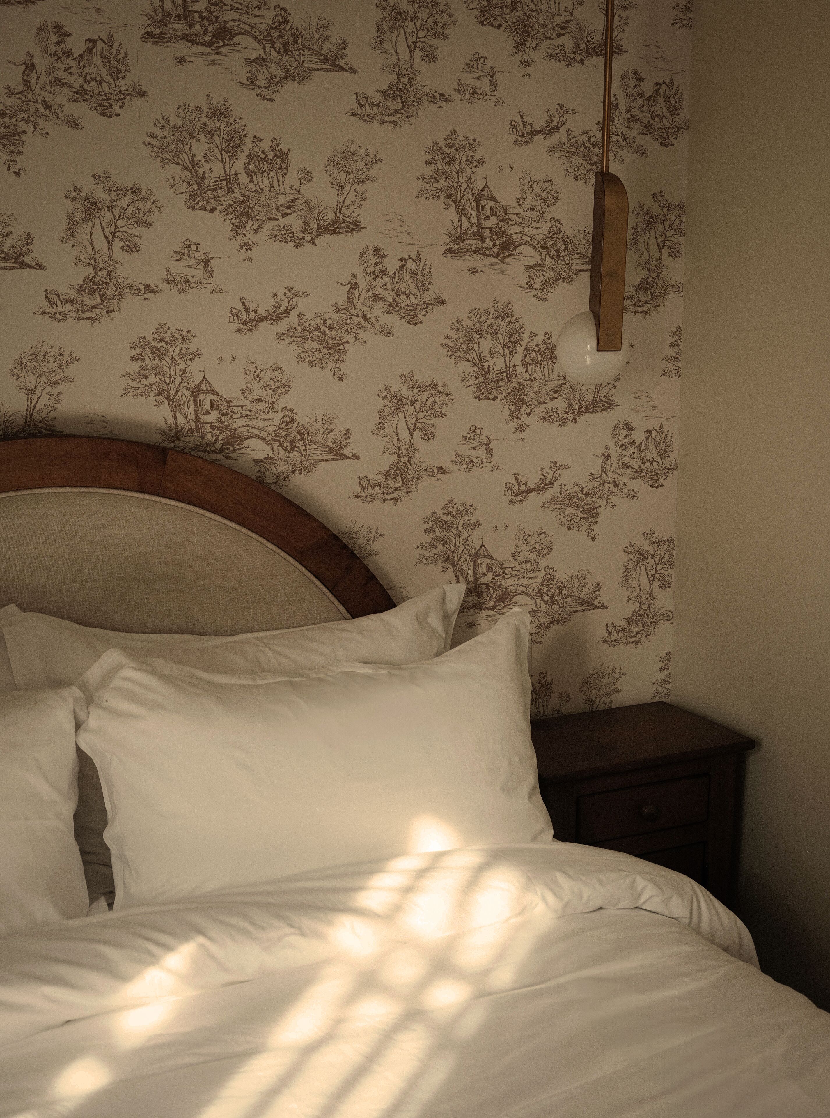 Un dormitorio con una cama, almohadas y una mesita de noche. La pared tiene un estampado de flores.