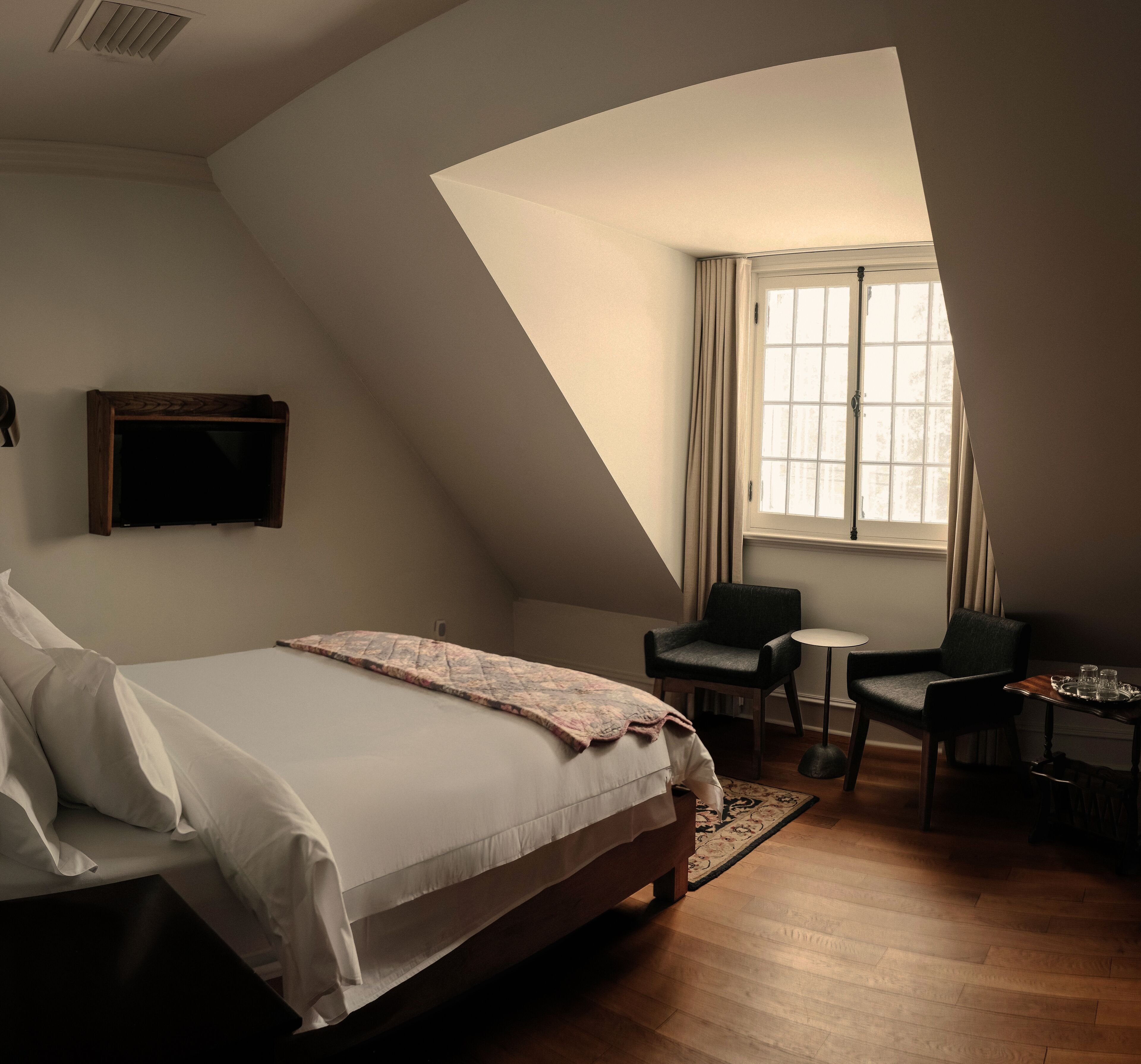 A bedroom with a large bed, a desk, two chairs, a small table, and a window with curtains.