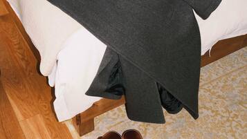 Una cama con un edredón blanco y una manta gris, un par de zapatos marrones y una chaqueta gris oscuro sobre la cama.