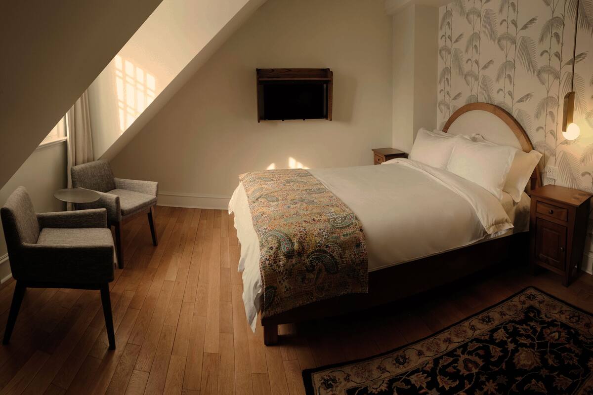 Une chambre à coucher avec un grand lit, une chaise, une petite table, un téléviseur fixé au mur et un tapis à motifs.