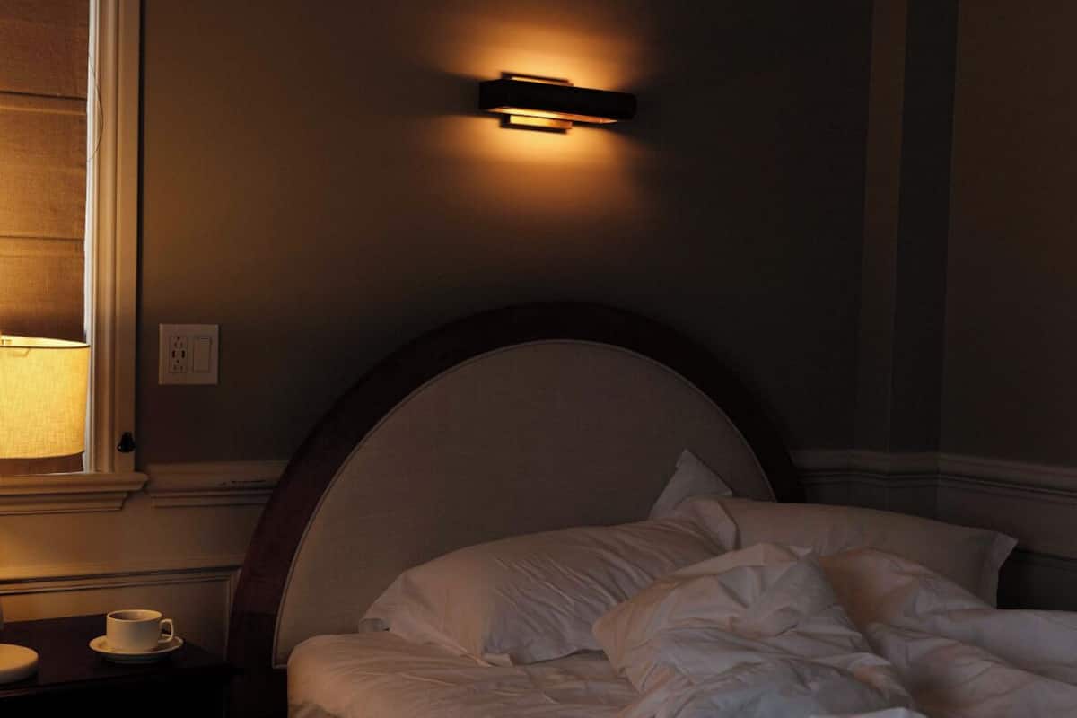 Une chambre à coucher avec un lit, une table de chevet sur laquelle se trouve une tasse, et une lampe fixée au mur.