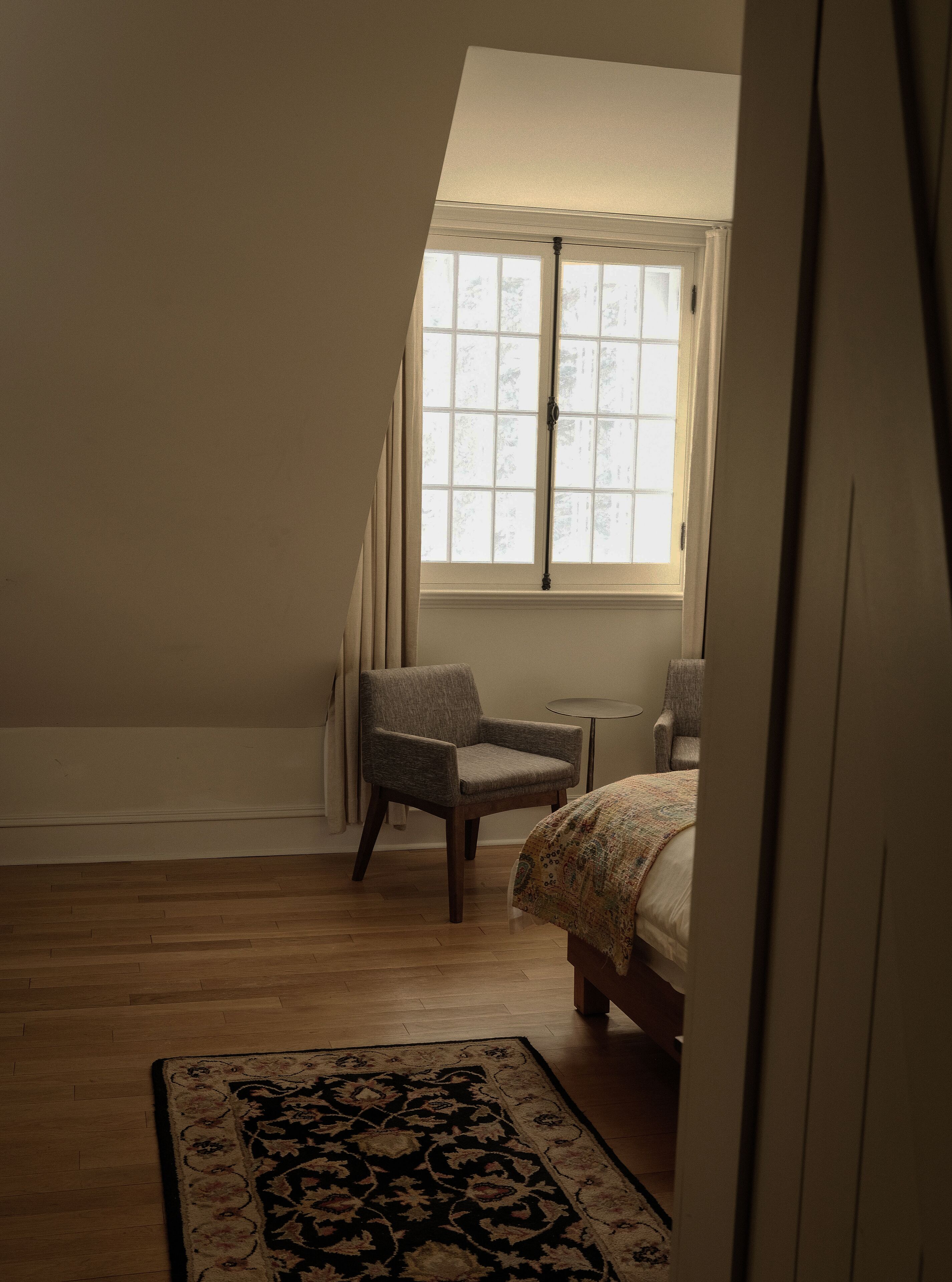 Une chambre avec un plafond en pente, une fenêtre avec des rideaux, un lit, une chaise et un tapis à motifs.