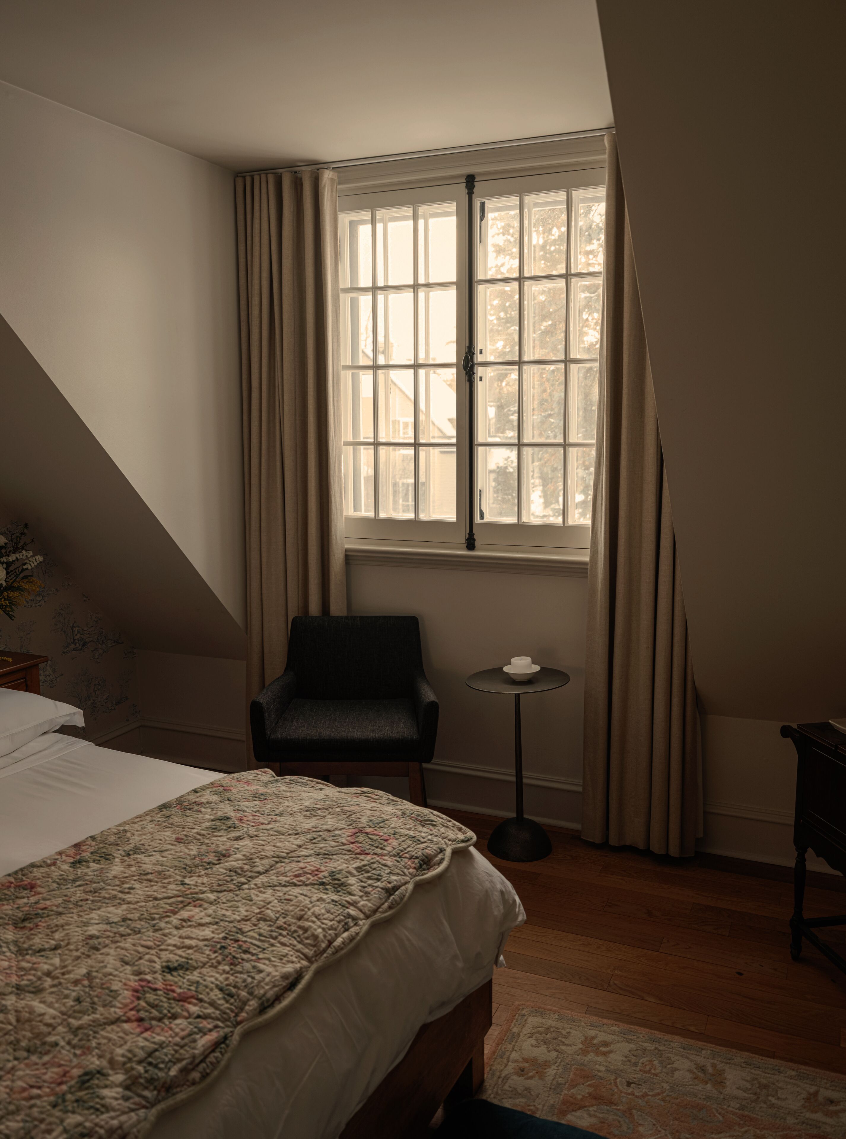Ein Schlafzimmer mit einem Bett, einem Sessel, einem kleinen Tisch und einem Fenster mit Vorhängen.