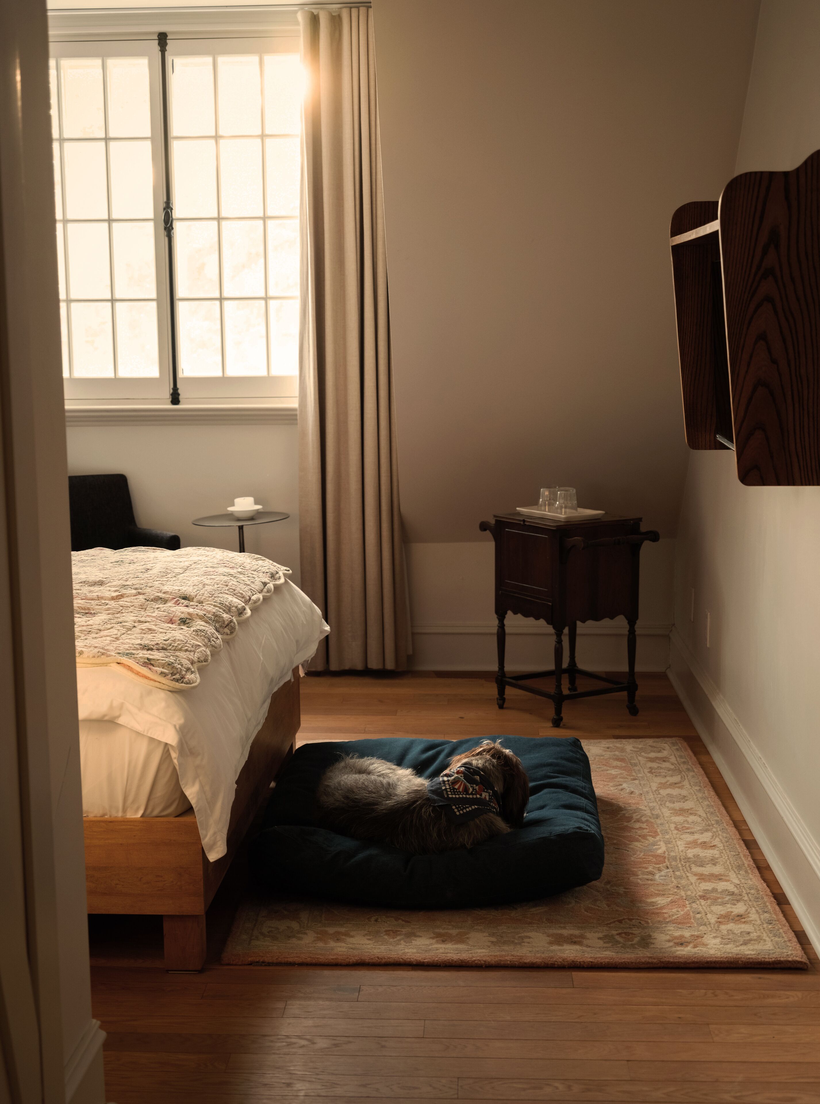 Ein Schlafzimmer mit einem Bett, einem kleinen Tisch, einem Stuhl, einem Hund auf einem Kissen, einem Fenster mit Vorhängen und Holzdielen.