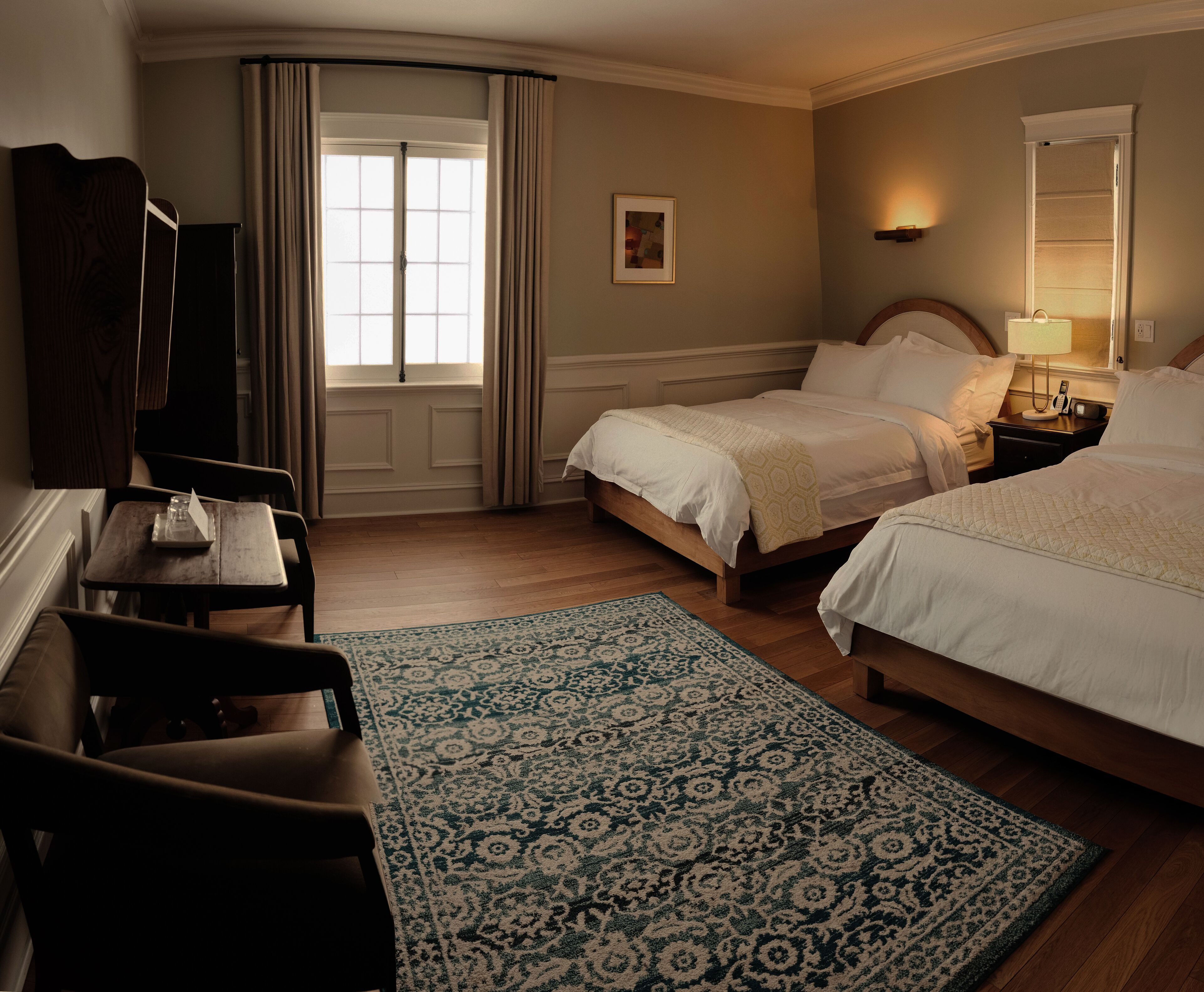 Une chambre d’hôtel avec deux lits, un bureau, une chaise, un tapis à motifs, une fenêtre avec des rideaux et un tableau encadré au mur.