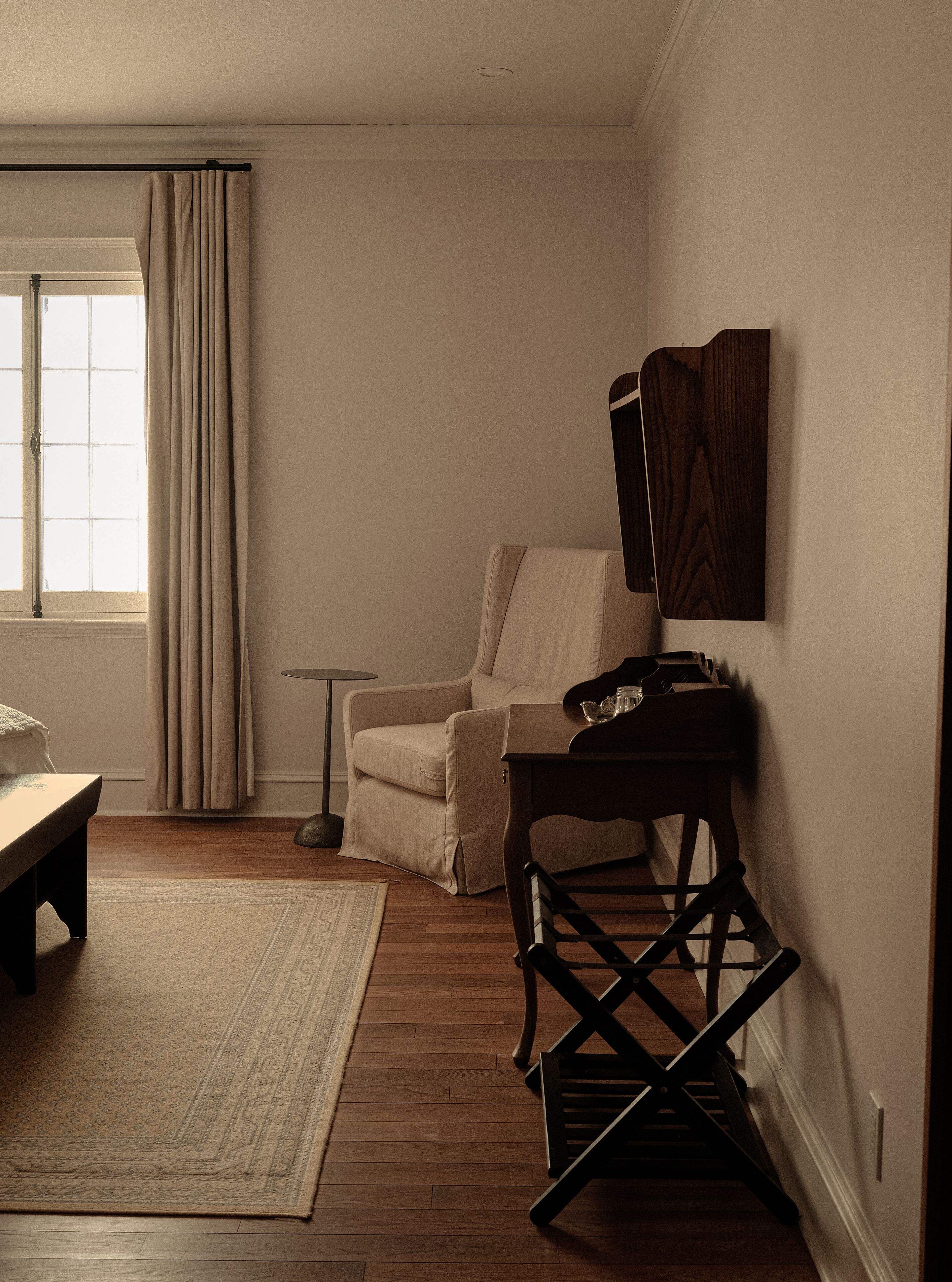 A room with a wooden floor, a beige armchair, a small table with a teapot, and a dark wooden chair.