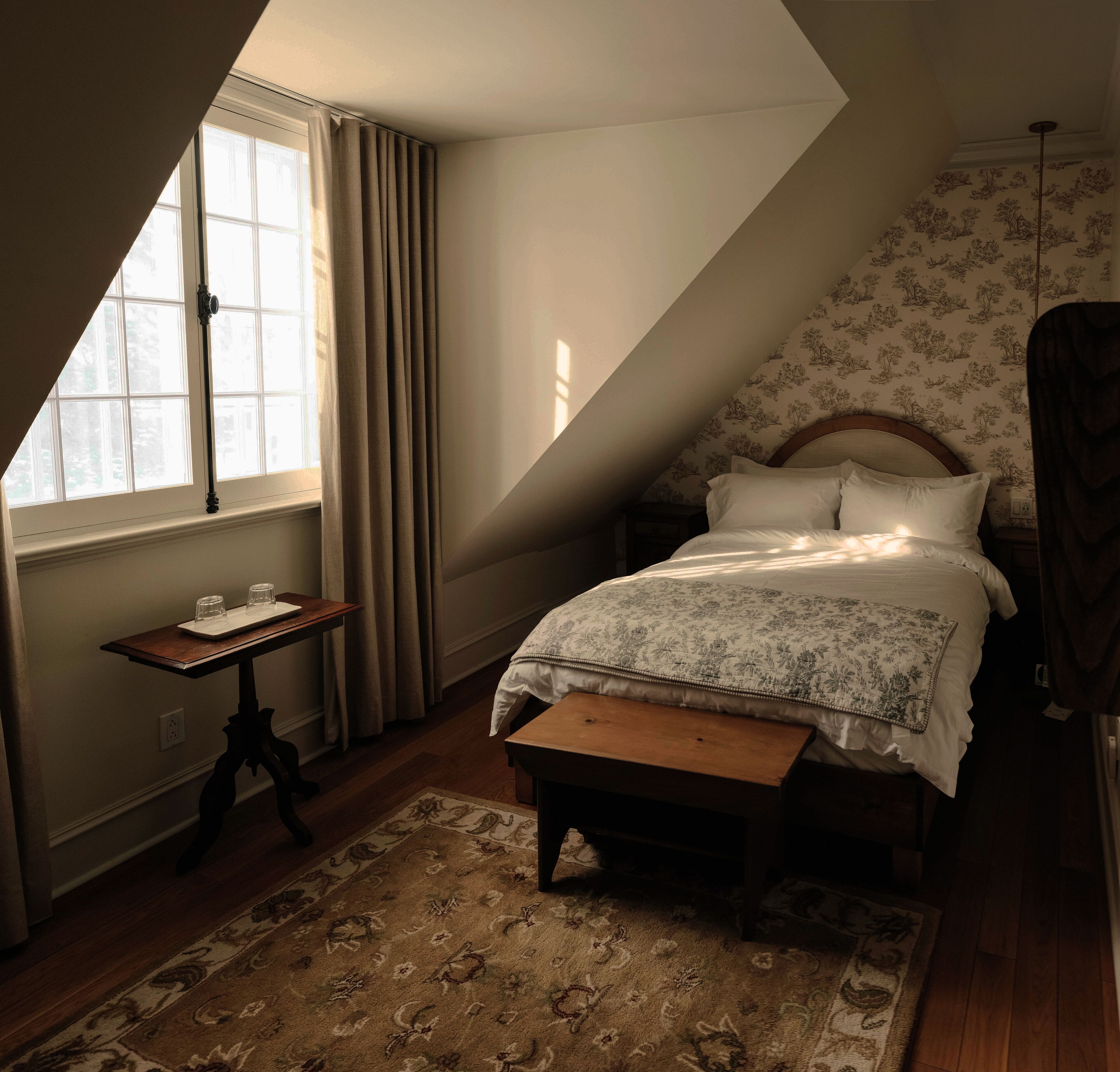 A bedroom with a sloped ceiling, a bed with floral bedding, a nightstand, a table with glasses, and a window with curtains.