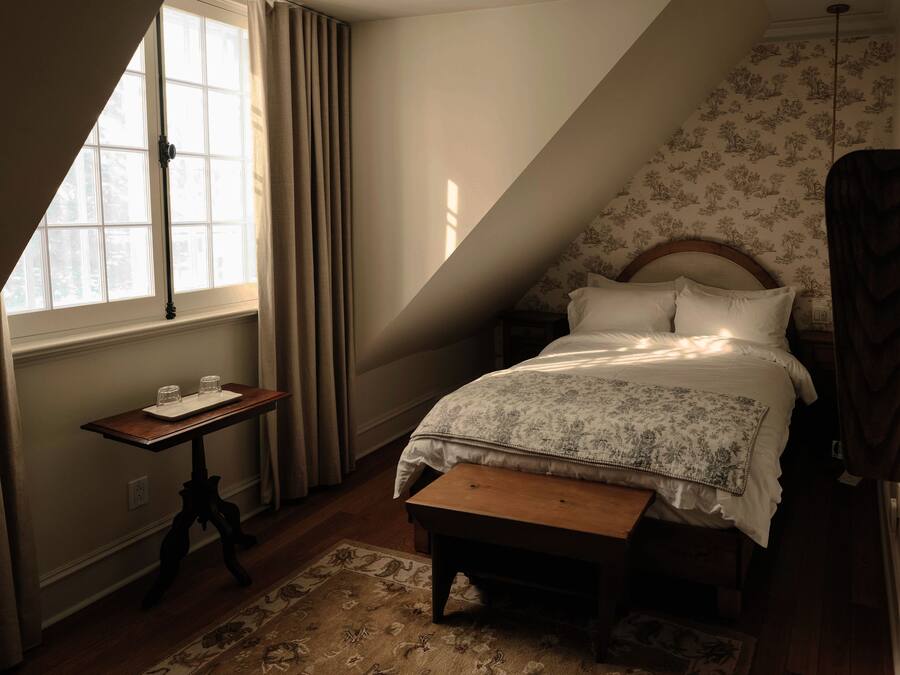 A bedroom with a sloped ceiling, a bed with floral bedding, a nightstand, a table with glasses, and a window with curtains.