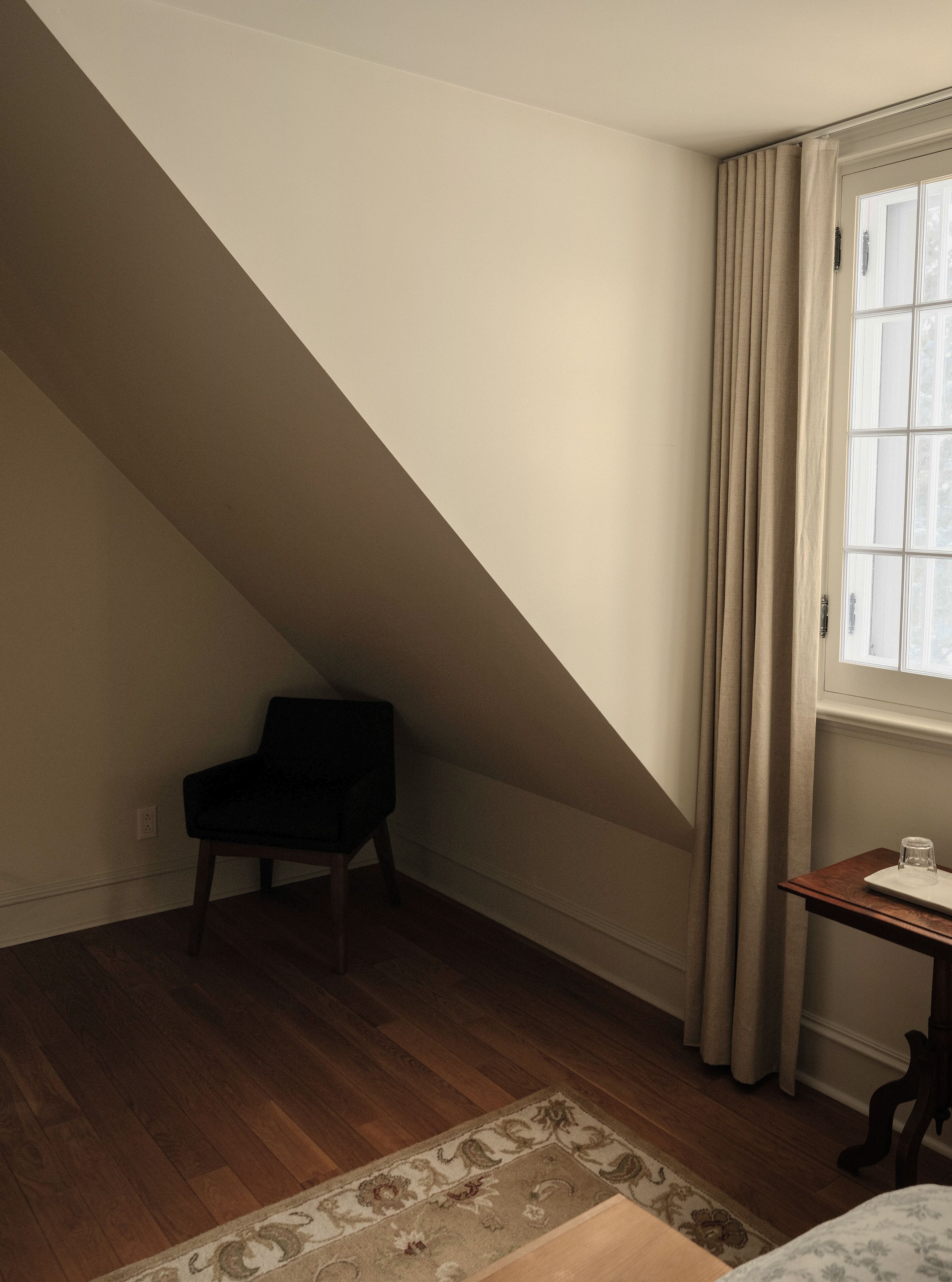 A room with a sloped ceiling, a chair, a small table with a glass, and a window with curtains.