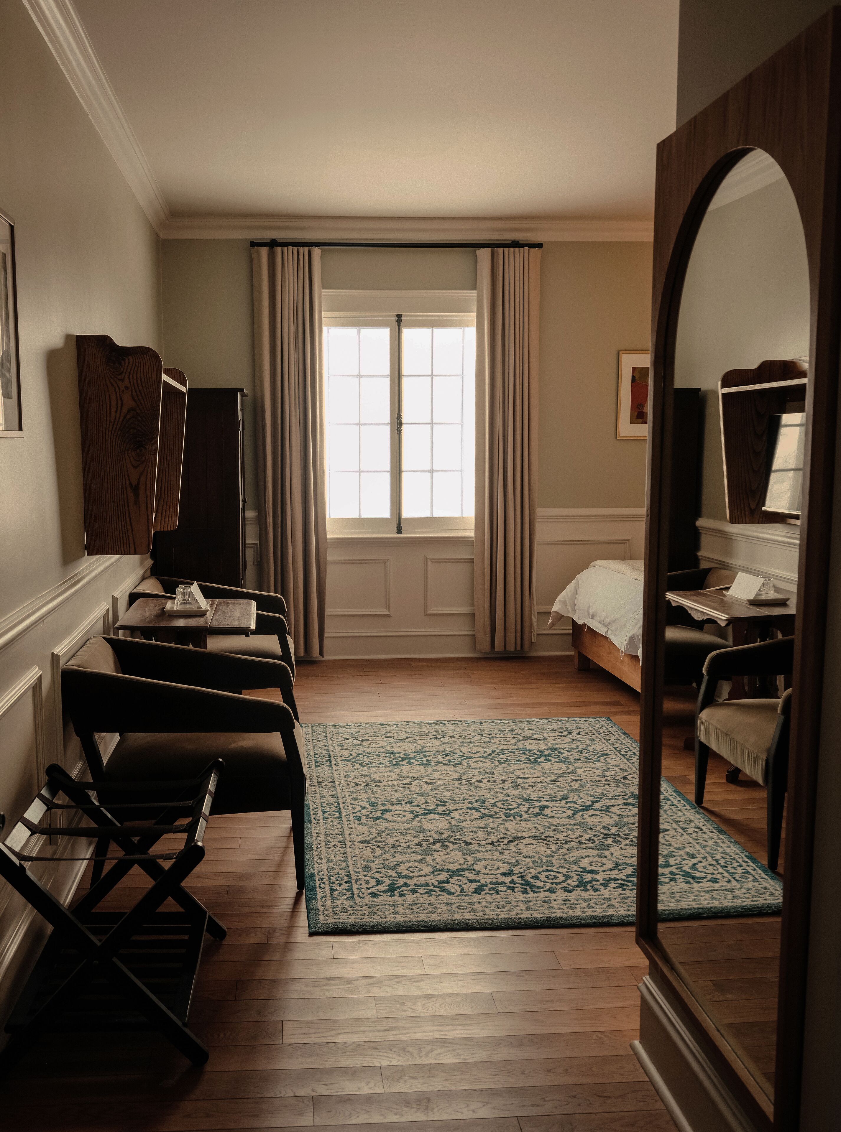 Una habitación de hotel con un ventanal grande, una cama, un escritorio con silla, un espejo y piso de madera.