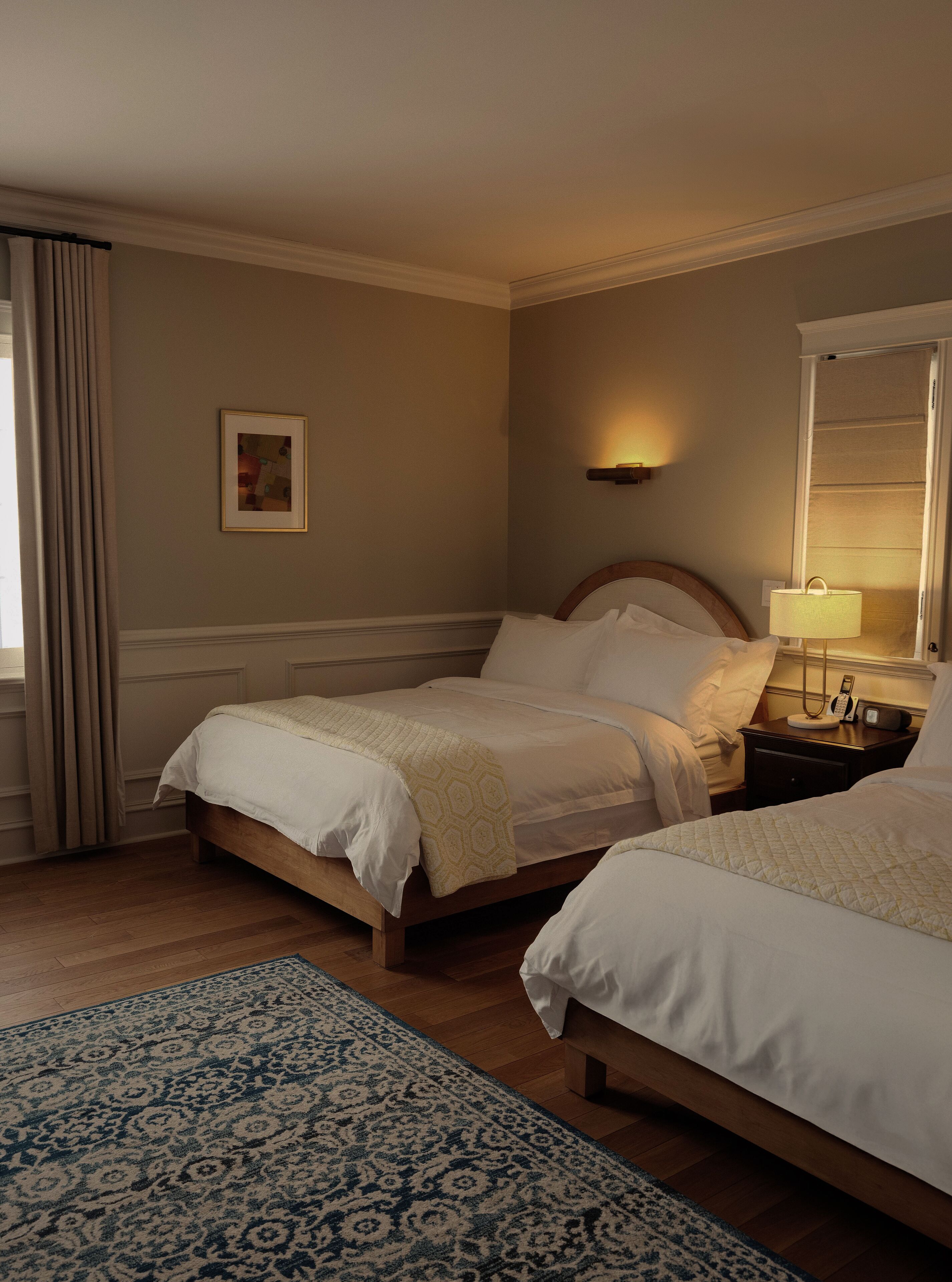 A hotel room with two beds, a bedside table with a lamp, a framed picture on the wall, and a patterned rug.