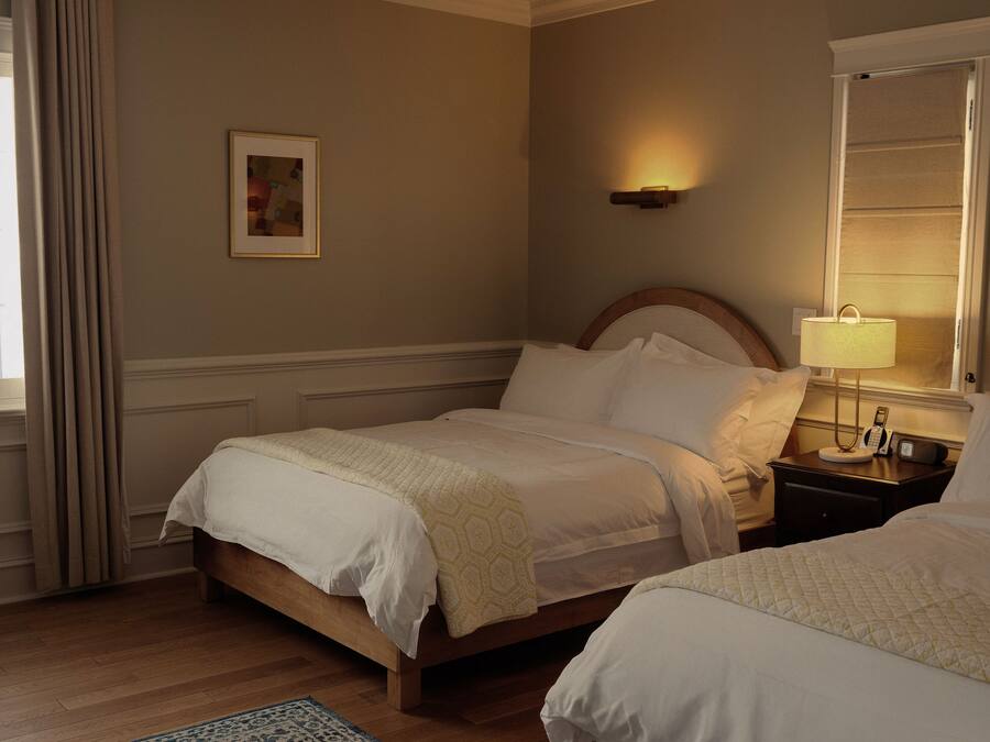 A hotel room with two beds, a bedside table with a lamp, a framed picture on the wall, and a patterned rug.