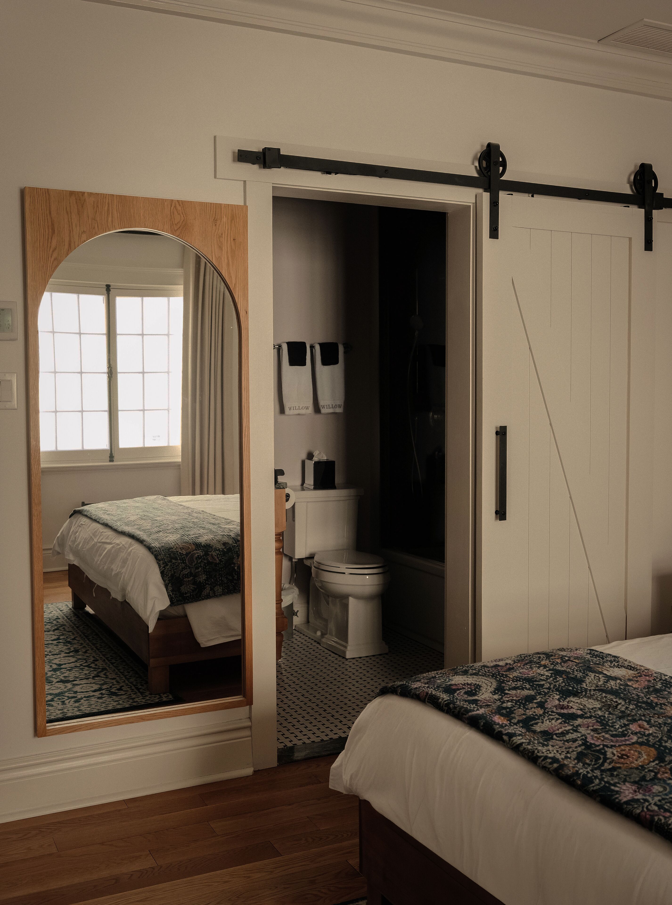 Une chambre à coucher comprenant un lit, un grand miroir, une armoire, une salle de bain avec des toilettes et une douche, ainsi qu’une fenêtre avec des rideaux.