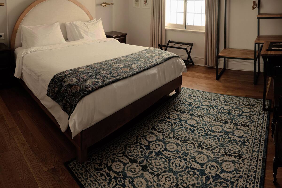 Une chambre à coucher bien rangée, avec un grand lit, une table de chevet en bois, un bureau et une fenêtre avec des rideaux.