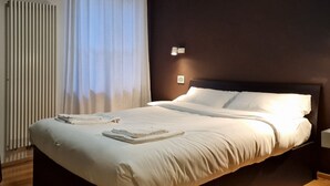 Quarto de hotel com cama, radiador, janela com cortinas e luminária fixada na parede.