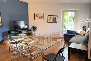 Een moderne woonkamer met een glazen eettafel, transparante stoelen, een bank en een televisie.