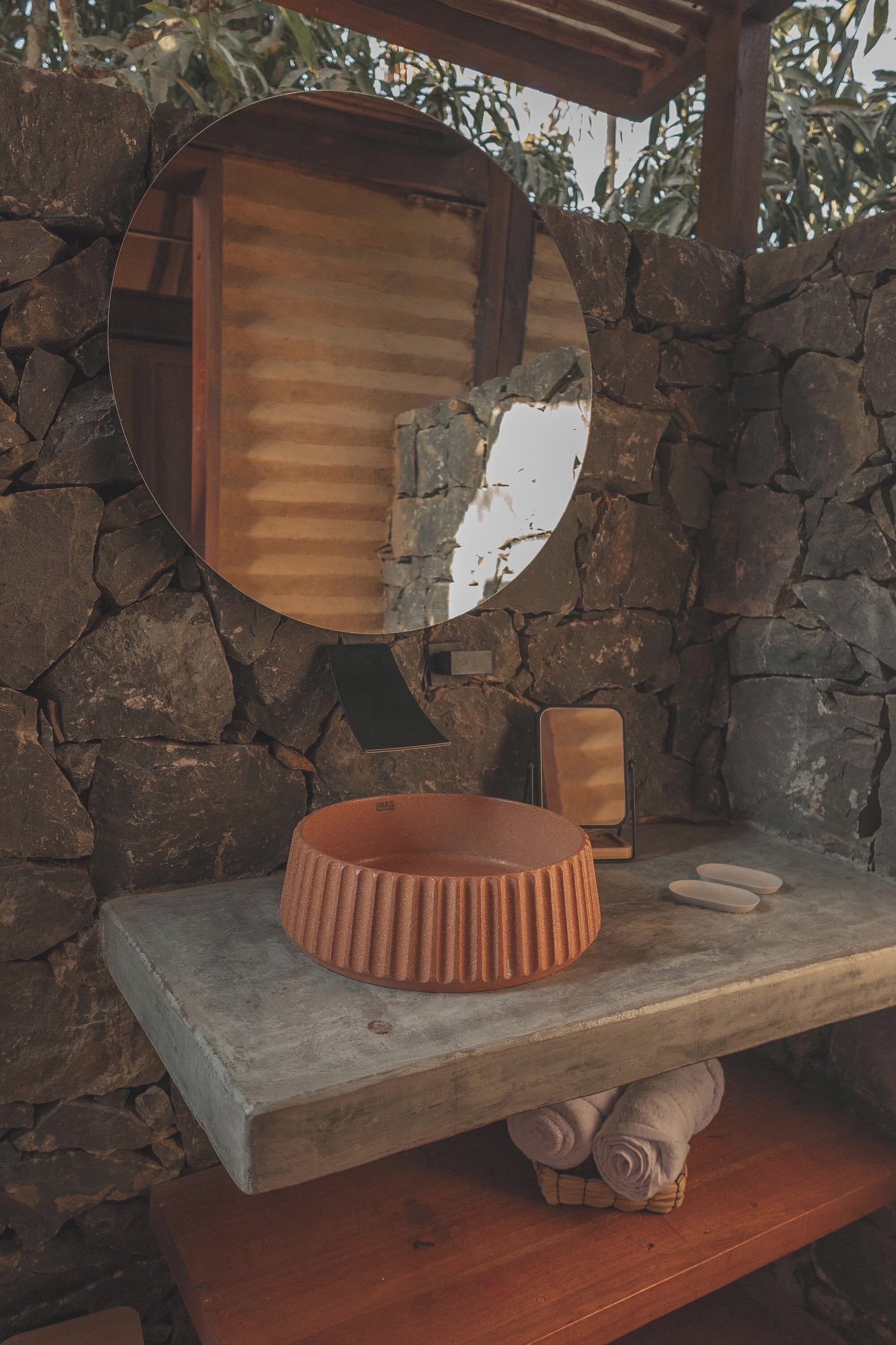 Pared de piedra con un espejo redondo, un banco de madera y un lavabo de piedra.