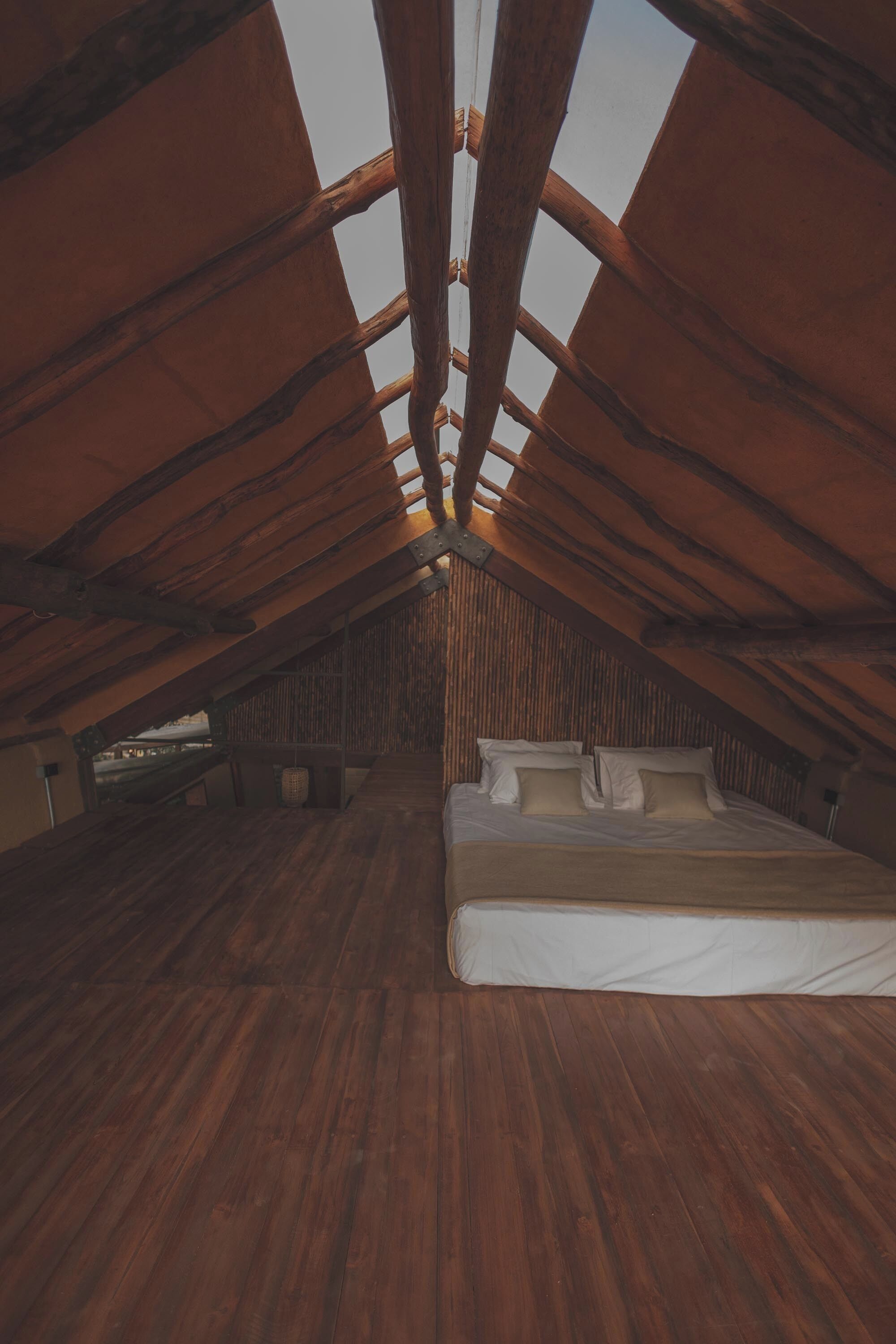 Une chambre mansardée avec un plancher en bois, un lit recouvert de draps blancs et un plafond en pente avec des poutres apparentes.