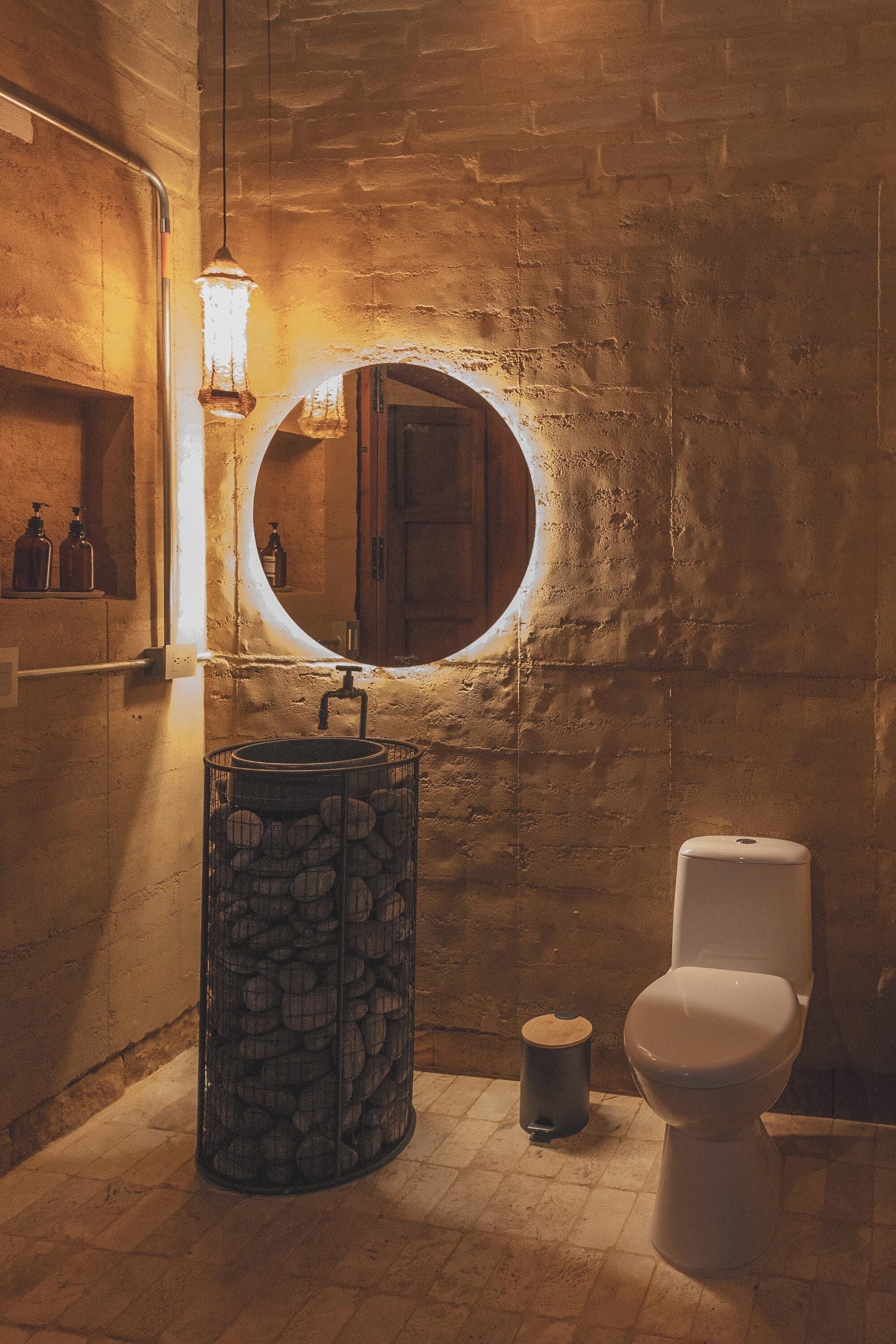 A bathroom with a rustic brick wall, a round mirror, a toilet, and a metal container with stones.