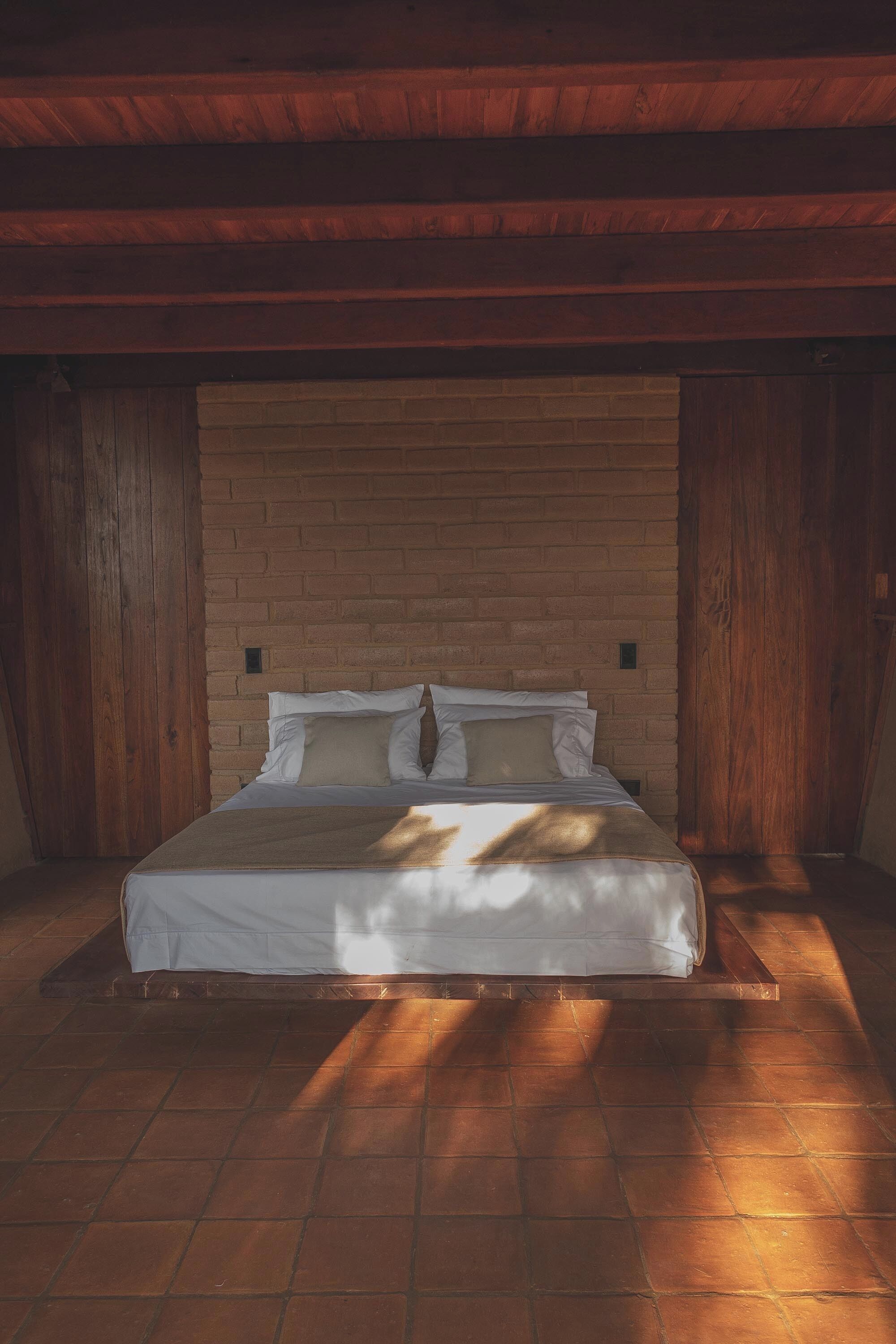 A bedroom with a brick wall, wooden ceiling beams, and a bed with white bedding.