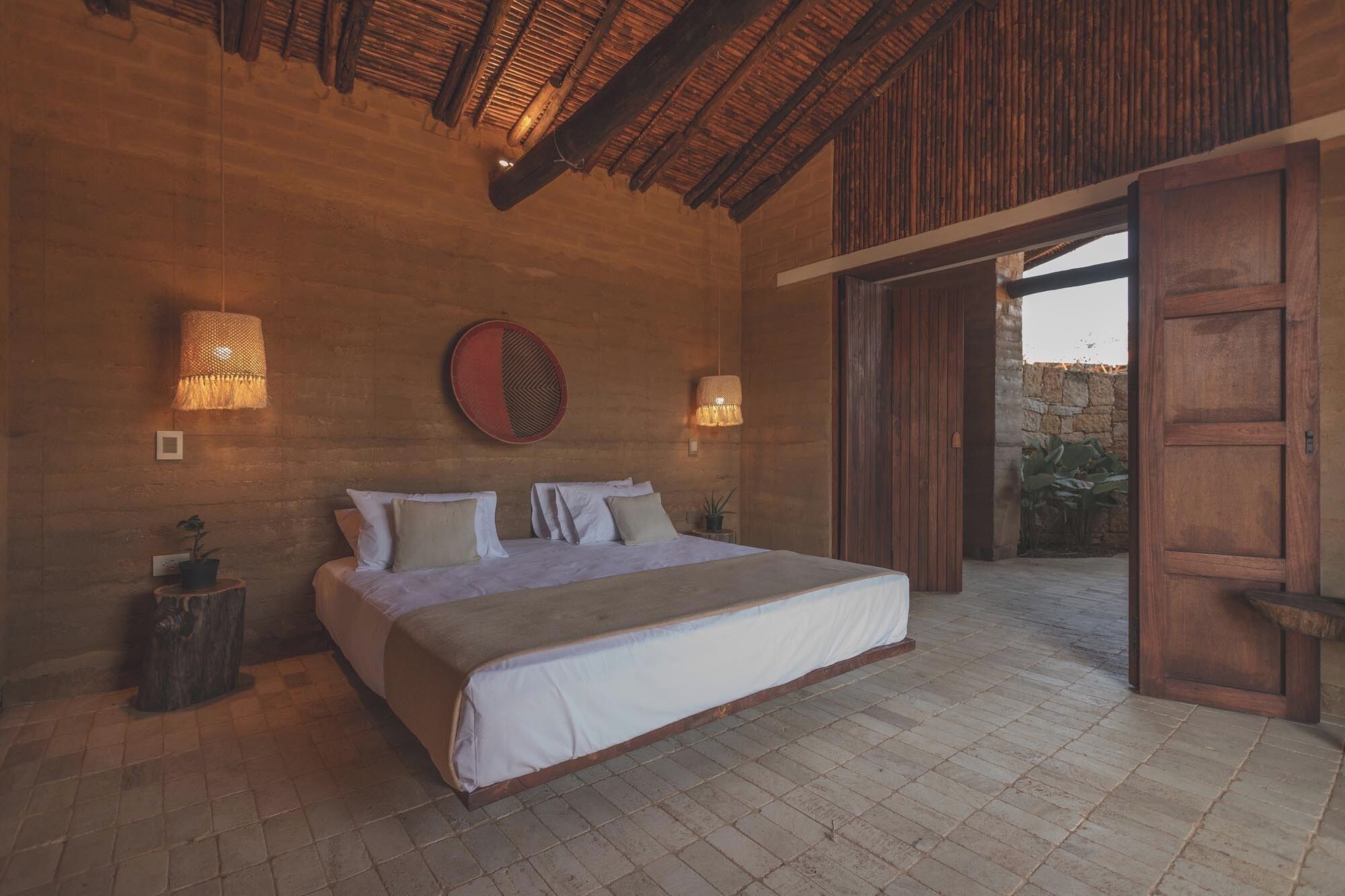 Une chambre spacieuse avec un grand lit, des poutres apparentes en bois au plafond et une vue sur un espace extérieur accessible par une porte ouverte.