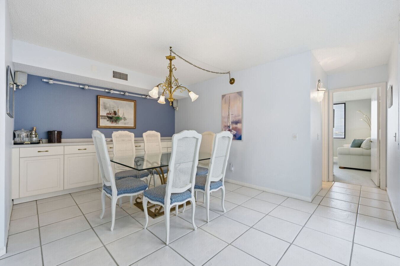 A dining area with a glass table and white chairs, a chandelier, and a painting on the wall.