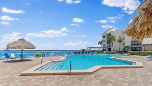 Ein Swimmingpool mit einer Strohdachschirm, blauen Liegestühlen und Blick auf das Meer.
