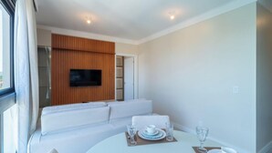 A modern room with a white sofa, a dining table with a set of plates and glasses, and a flat-screen TV mounted on a wooden panel wall. - Fully Equipped apt Near the Beach Rcq0305 (Florianópolis)