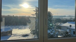 Blick durch ein Fenster auf eine verschneite Landschaft mit Bäumen und einem Gebäude mit Schnee bedecktem Dach.