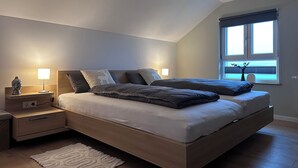 Ein modernes Schlafzimmer mit Schlafplatz in erhöhter Lage, Nachttischlampen, einer kleinen Statue und einem Fenster mit Jalousien.