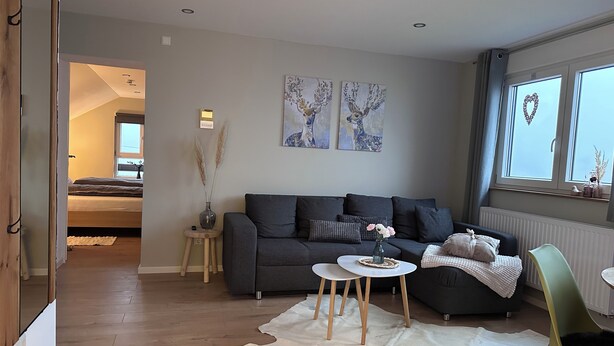 Ein modernes Wohnzimmer mit einem grauen Sofa, einem weißen Teppich und einem kleinen Tisch mit einer Vase. An der Wand hängen zwei Gemälde und ein Fenster mit Blick.