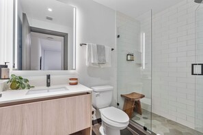 Une salle de bain moderne avec un lavabo blanc, des toilettes, une cabine de douche en verre et un petit tabouret en bois.
