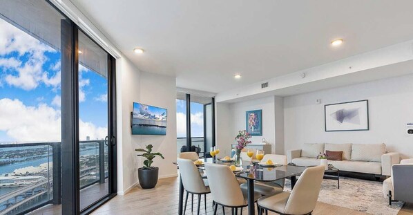 Un salon moderne avec un coin repas, de grandes fenêtres offrant une vue sur la ville et un téléviseur à écran plat fixé au mur.