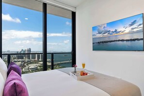 Une chambre d’hôtel dotée d’une grande fenêtre donnant sur la ville, d’un lit avec des oreillers, d’un plateau avec des fruits et une boisson, et d’un tableau représentant un paysage côtier.