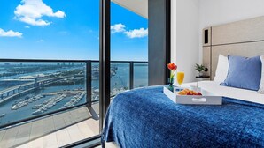 Quarto de hotel com uma varanda ampla, cama com colcha azul, bandeja com croissants e suco de laranja, e vista para um horizonte urbano com uma ponte e marina.