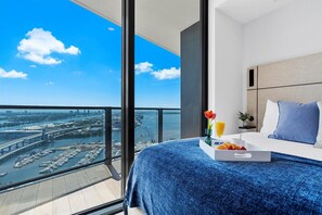 Ein Hotelzimmer mit großem Balkon, einem Bett mit blauer Bettdecke, einem Tablett mit Croissants und Orangensaft sowie Blick auf eine Stadtlandschaft mit Brücke und Yachthafen.
