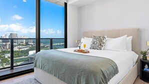 Ein modernes Schlafzimmer mit großem Fenster, einem Bett mit grauer Decke, weißen Kissen und einem Tablett mit Frühstücksgebäck.