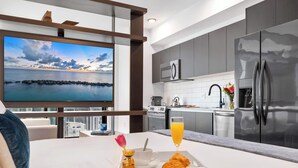A modern hotel room with a large TV displaying a scenic view, a breakfast tray with croissants, a glass of orange juice, and a vase with flowers. - Floor-to-Ceiling Views | Downtown Studio Suite (Miami)