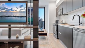 A modern kitchen with stainless steel appliances, a built-in oven, and a dishwasher. There is a wooden dining table with a white chair and a small decorative globe. A television is mounted on a wooden frame, displaying a scenic view. - Floor-to-Ceiling Views | Downtown Studio Suite (Miami)