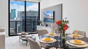 Un espace repas moderne avec vue sur la ville, une grande fenêtre et une table dressée pour deux personnes, avec des assiettes de nourriture et des verres.