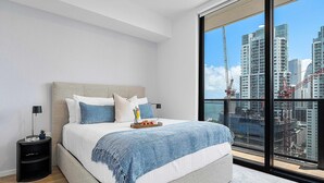 Une chambre moderne avec un grand lit, une table de chevet, une lampe et une vue sur le paysage urbain, visible à travers une grande fenêtre.