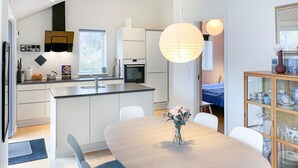 Een moderne keuken met witte kasten, een inbouwoven en een groot raam. Er is een eethoek met een houten tafel en stoelen, en een vaas met bloemen op de tafel.