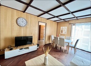 Una sala de estar moderna con techo de madera, televisor de pantalla plana, zona de comedor con mesa y sillas, y un ventanal que proporciona luz natural.